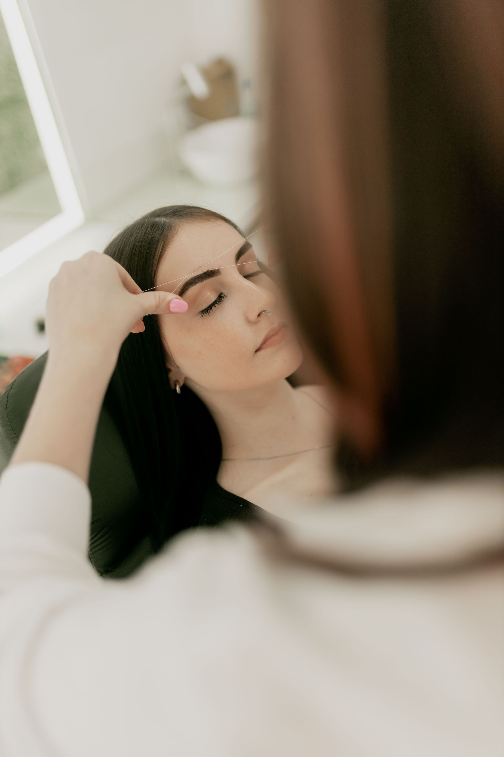A beautician shaping a young woman's eyebrows in a serene salon setting.